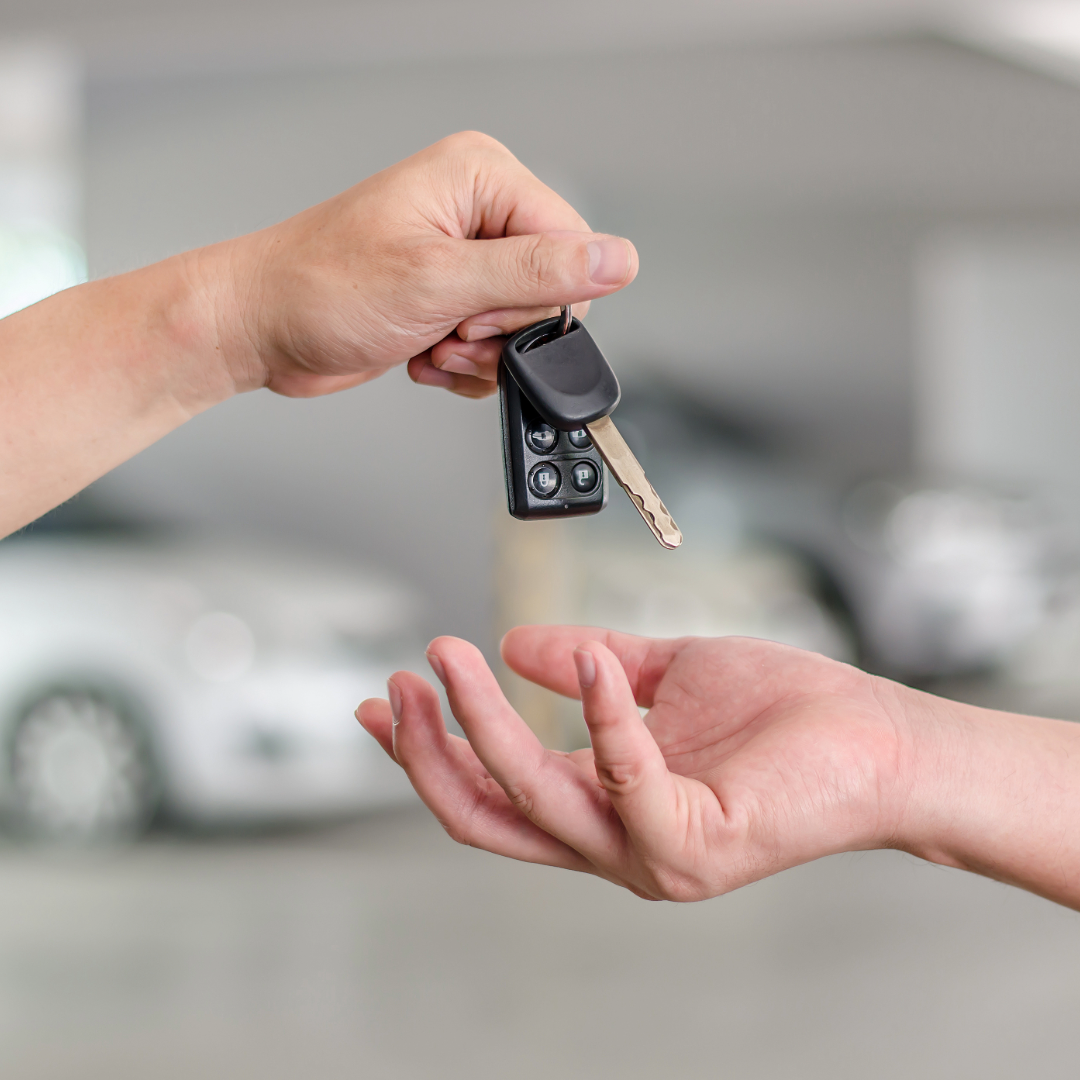 Hand holding out keys to another hand, with a car in the background.